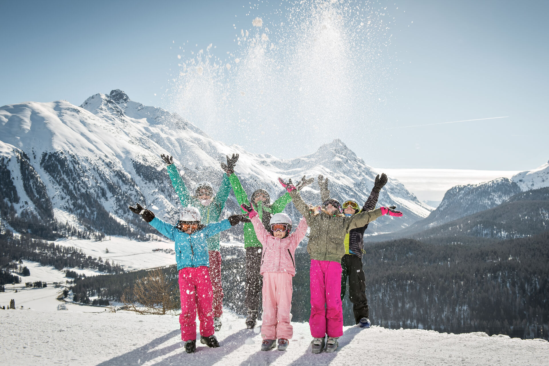 Sechs Kinder in verschiedenfarbener Skiausrüstung stehen auf einer Skipiste und werfen Schnee in die Luft, der auf sie zurückfällt. Im Hintergrund sind Waldflächen und das sich öffnende Oberengadiner Tal zu sehen, dahinter schneebedeckte Berge. Der Himmel ist wolkenlos und strahlend blau.