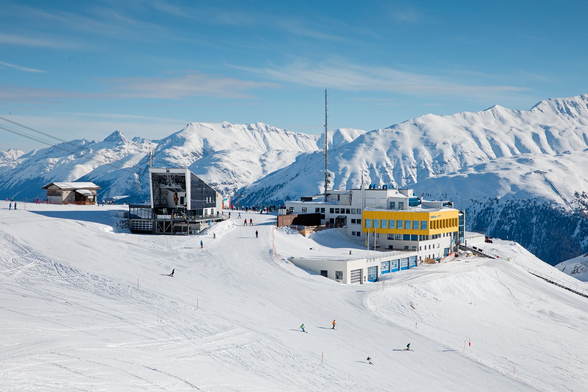 Verschneite Skigebietsszene auf Corviglia oberhalb von St. Moritz im Engadin. Mehrere Skifahrer sind auf den Skipisten unterwegs. Im Hintergrund sind die Bergstation Corviglia und die Talstation Piz Nair in einer winterlichen Berglandschaft zu sehen.