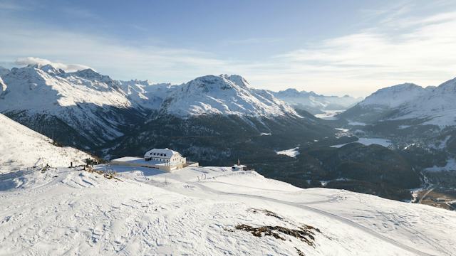 Aufnahme verschneiten Winterlandschaft auf Muottas Muragl im Engadin. Das Romantik Hotel steht auf einem schneebedeckten Hang, dahinter erstrecken sich schneebedeckte Alpen, Täler und gefrorene Seen unter blauem Himmel.