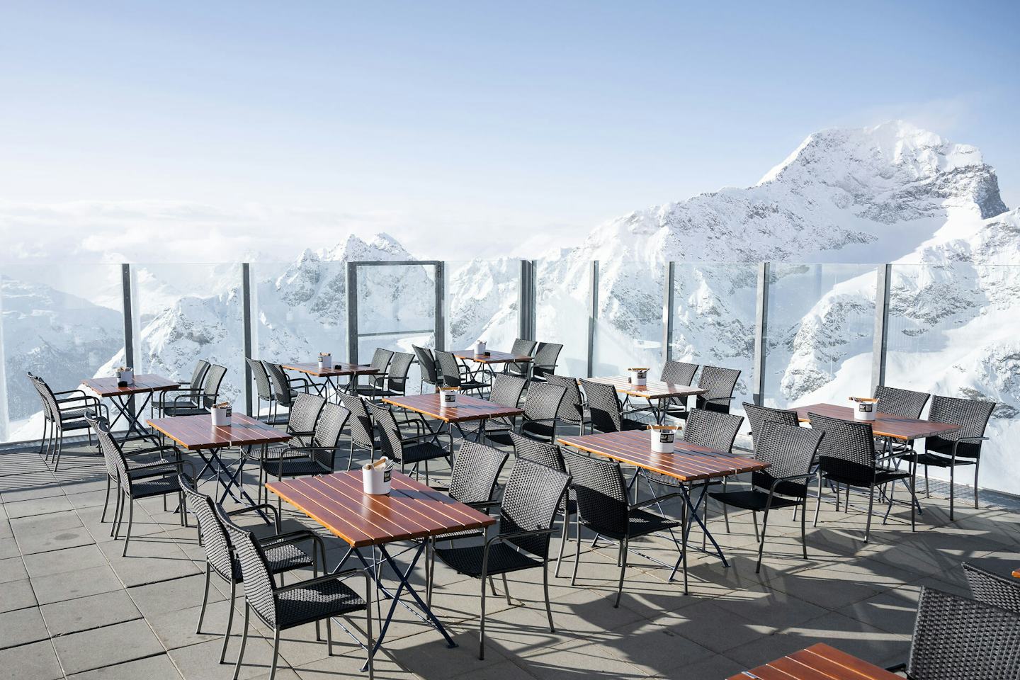 Die Panoramaterrasse des Bergrestaurants Piz Nair 10000 Feet auf 3057 Metern. In der Sonne stehen gedeckte Tische mit Blick auf die verschneite Alpenlandschaft rund um den St. Moritzer Hausberg Piz Nair. Im Hintergrund sind Berge mit weissen Gipfeln und Berghängen sowie ein weiter, blauer und wolkenloser Himmel zu sehen.