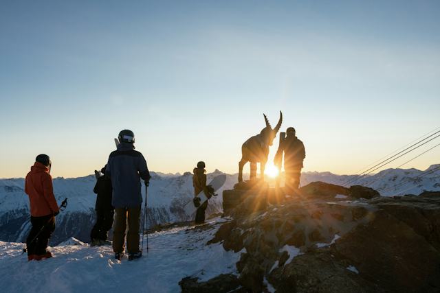 Piz Nair Sunrise Winter Winterlandschaft auf dem Piz Nair bei St. Moritz im Engadin während des Sonnenaufgangs. Die Sonne geht zwischen einer Steinbock-Statue und einem Skifahrer auf. Zwei weitere Skifahrer und zwei Snowboarder beobachten den spektakulären Sonnenaufgang ebenfalls. Verschneite Berge und Hänge werden von warmem Morgenlicht beleuchtet.