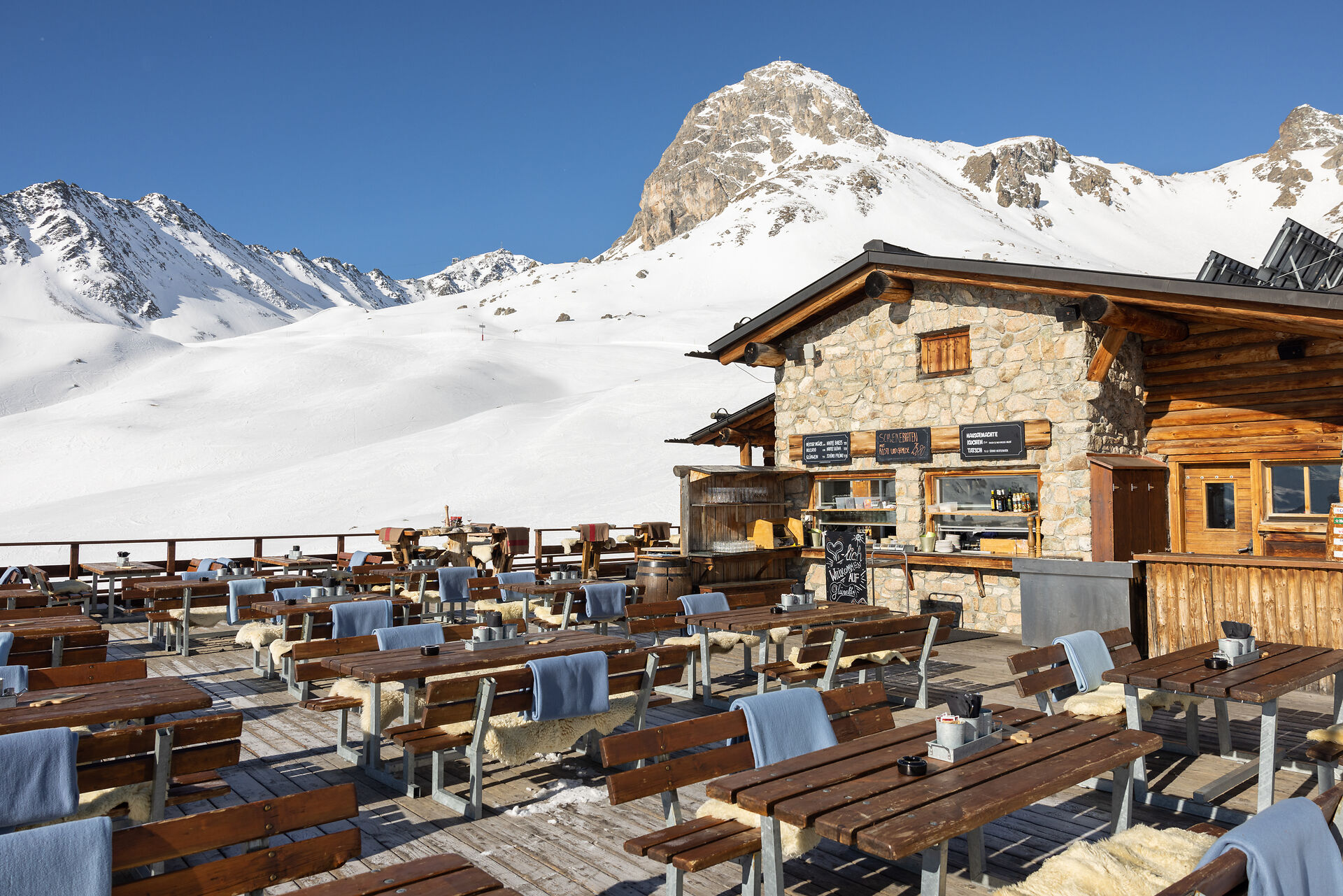Aussenansicht der Sonnenterrasse des Restaurants Glünetta in Marguns. Vor dem Gebäude stehen Holztische und Sitzbänke. Dahinter sind schneebedeckte Skipisten des Skigebiets Corviglia sowie ein klarer, wolkenloser blauer Himmel zu sehen.