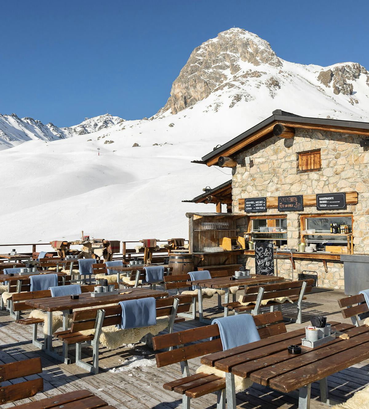 Restaurant Glünetta auf Marguns im Skigebiet Corviglia | Winter Aussenansicht der Sonnenterrasse des Restaurants Glünetta in Marguns. Vor dem Gebäude stehen Holztische und Sitzbänke. Dahinter sind schneebedeckte Skipisten des Skigebiets Corviglia sowie ein klarer, wolkenloser blauer Himmel zu sehen.
