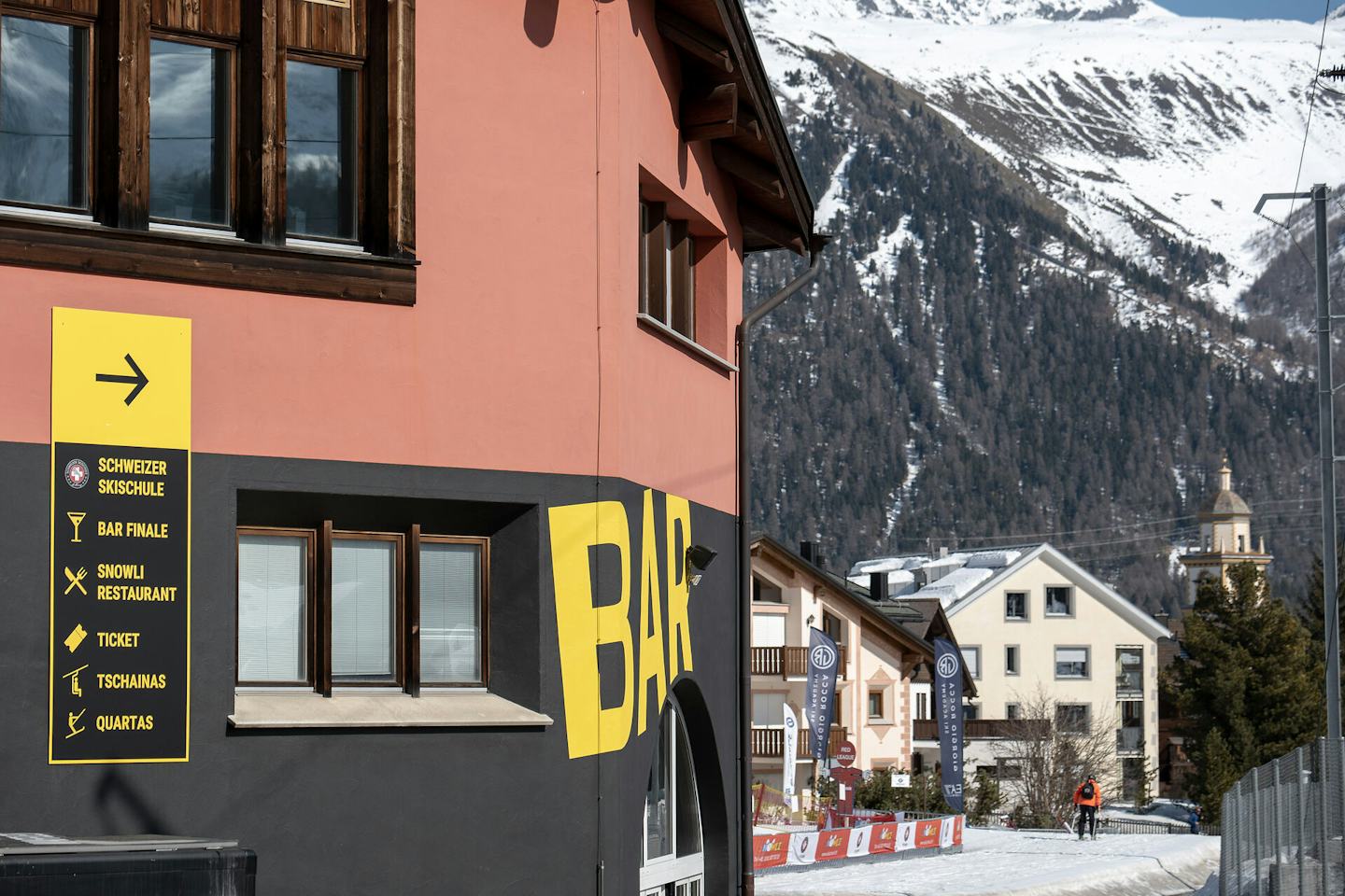 Aussenansicht der Bar Finale in Celerina im Engadin. Ein modernes Gebäude mit orangefarbener Fassade und grossen Fenstern steht an den Bahngleisen der Rhätischen Bahn. Im Hintergrund sind die Ortschaft Celerina, ein Kirchturm, weisse Berghänge und die alpine Landschaft zu sehen.