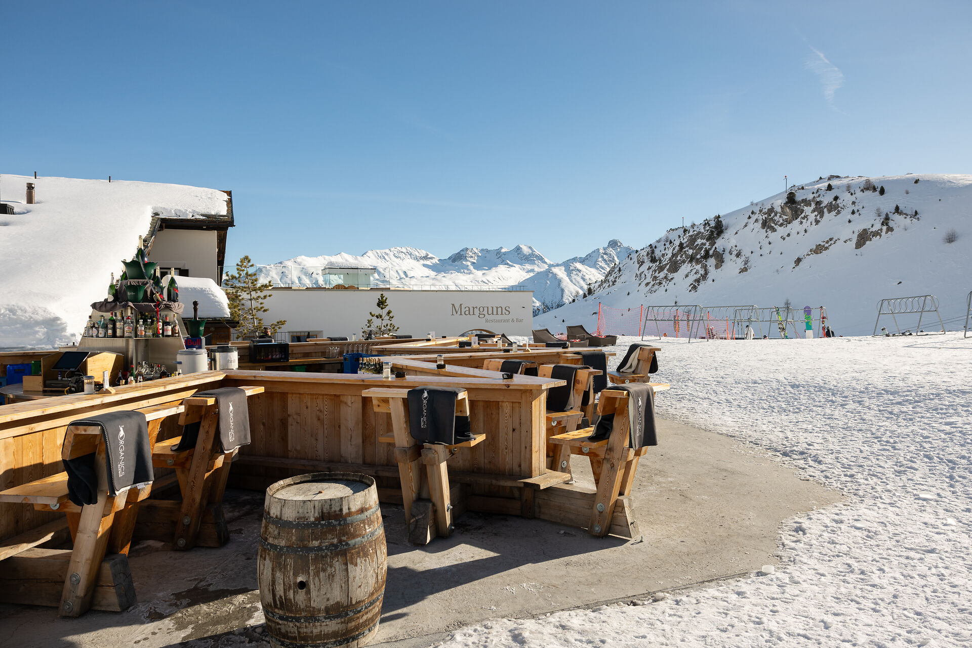 Das Sternbar Marguns präsentiert sich als sternförmige Bar direkt an der Skipiste auf dem Marguns. An der Holzbar stehen Barhocker aus Holz und Fässer, die als Stehtische um die Bar herum platziert sind. Im Hintergrund sind das Gebäude des Restaurants Marguns, Skiständer und die weissen Hänge der Berglandschaft des Oberengadins zu sehen – und das alles bei blauem, wolkenlosem Himmel.