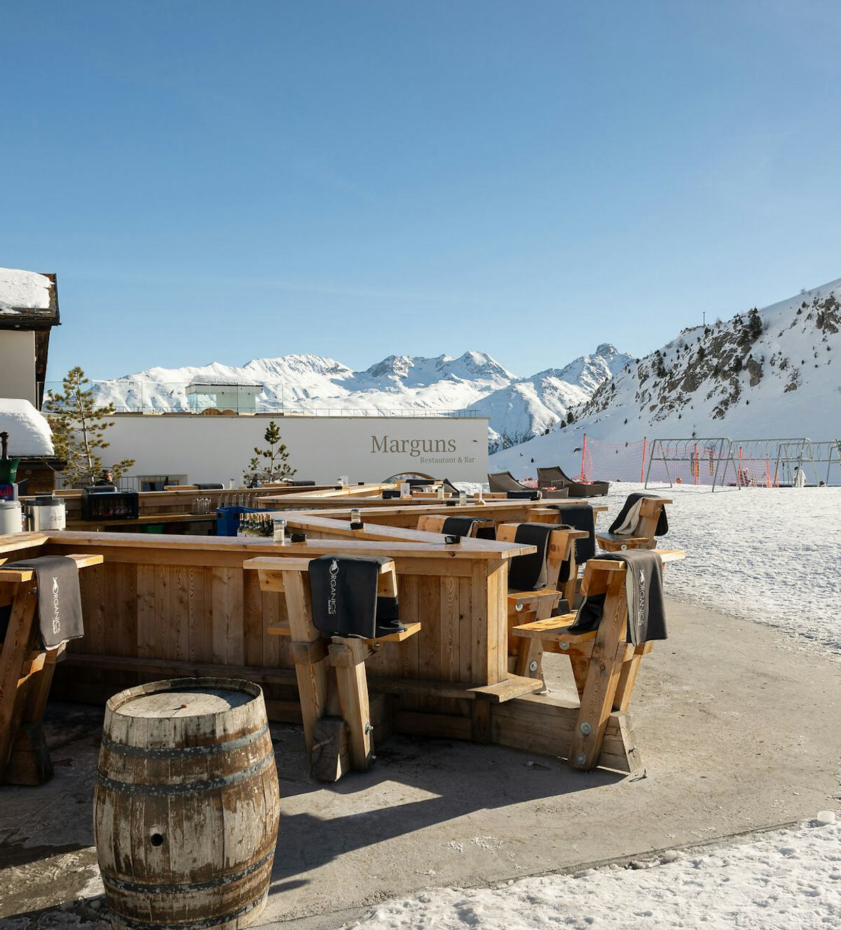 Sternbar Marguns | Winter Das Sternbar Marguns präsentiert sich als sternförmige Bar direkt an der Skipiste auf dem Marguns. An der Holzbar stehen Barhocker aus Holz und Fässer, die als Stehtische um die Bar herum platziert sind. Im Hintergrund sind das Gebäude des Restaurants Marguns, Skiständer und die weissen Hänge der Berglandschaft des Oberengadins zu sehen – und das alles bei blauem, wolkenlosem Himmel.