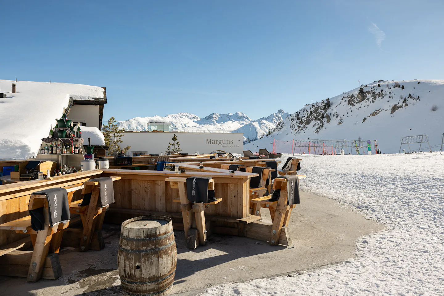 Sternbar Marguns | Winter Das Sternbar Marguns präsentiert sich als sternförmige Bar direkt an der Skipiste auf dem Marguns. An der Holzbar stehen Barhocker aus Holz und Fässer, die als Stehtische um die Bar herum platziert sind. Im Hintergrund sind das Gebäude des Restaurants Marguns, Skiständer und die weissen Hänge der Berglandschaft des Oberengadins zu sehen – und das alles bei blauem, wolkenlosem Himmel.