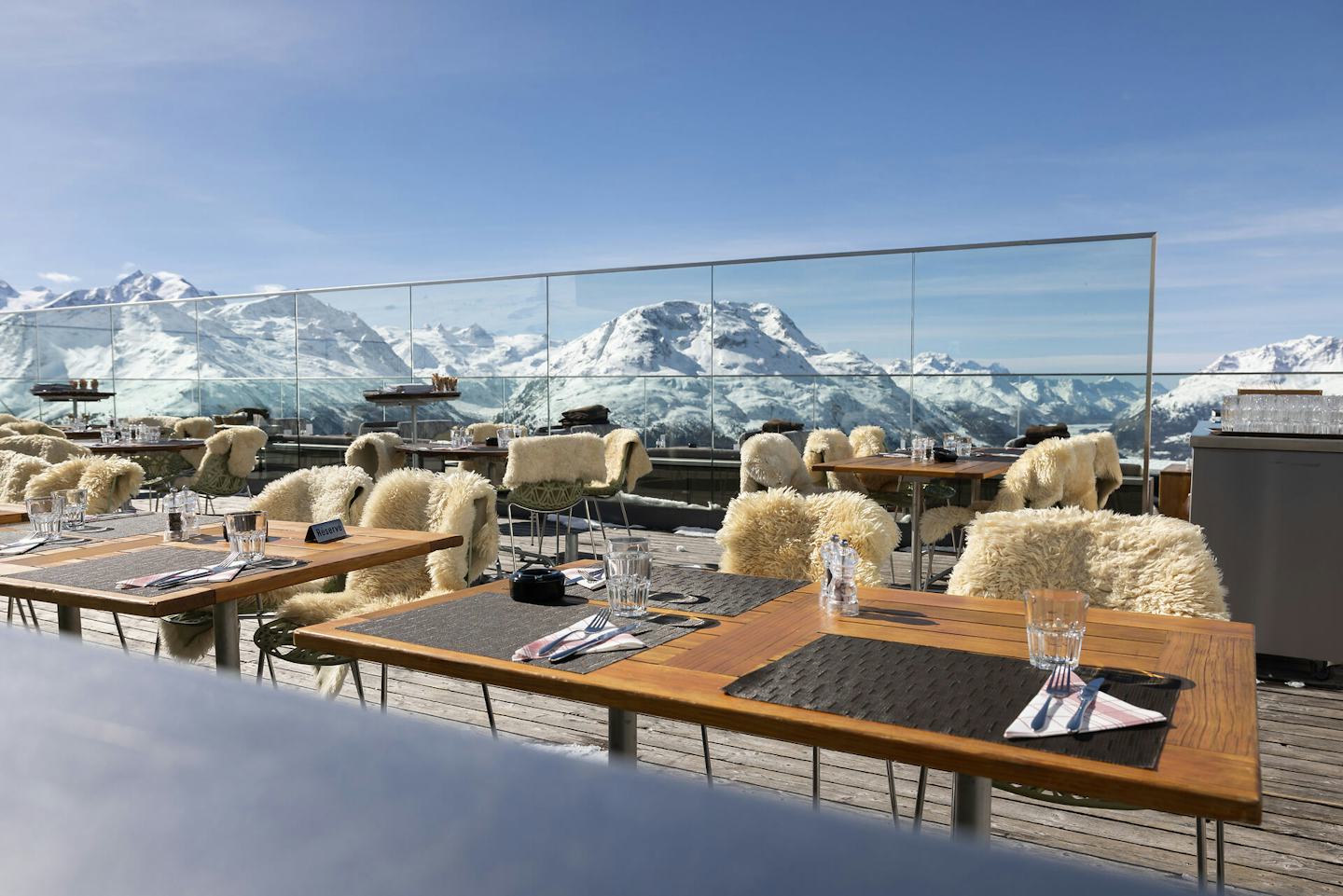 Panoramaterrasse | Romantik Hotel Muottas Muragl | Winter Die grosszügige Panoramaterrasse des Romantik Hotels Muottas Muragl bietet zahlreiche Sitzplätze im Freien. Sie ist mit Tischen, Stühlen und Lammfellen ausgestattet und liegt an einem Hang. Im Hintergrund erstreckt sich eine weitläufige, schneebedeckte Berglandschaft mit Tälern und Gipfeln unter klarem Himmel.