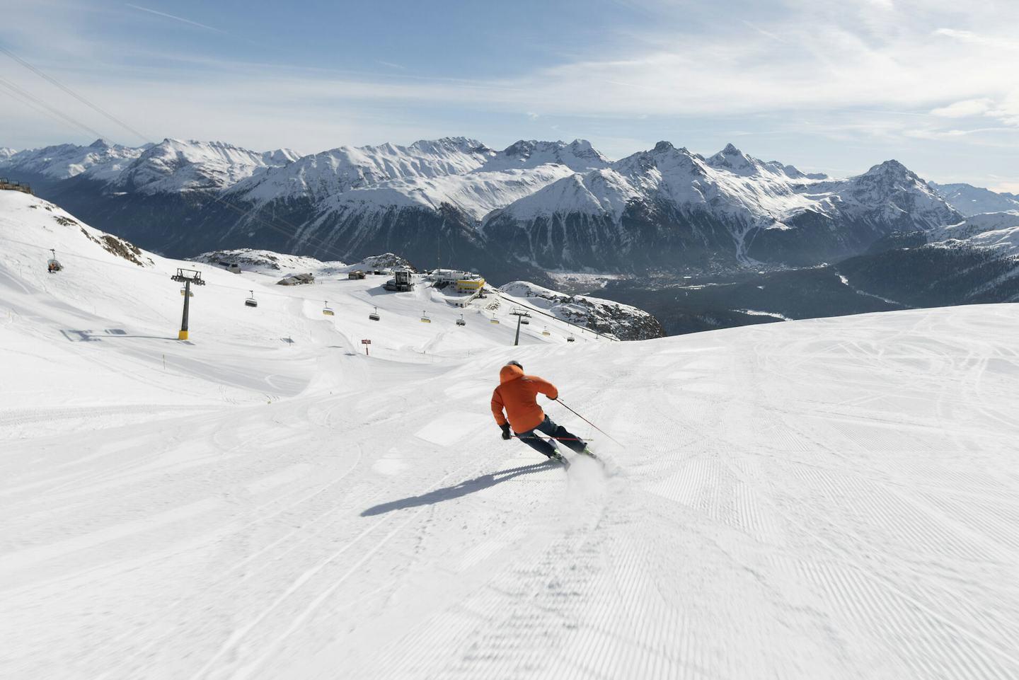 Ein einzelner Skifahrer carvt auf perfekt präparierten Pisten in Richtung Bergstation Corviglia. Vor ihm öffnet sich das schneebedeckte Tal mit schneebedeckten Gipfeln. Links im Bild ist eine Sesselbahn zu sehen.