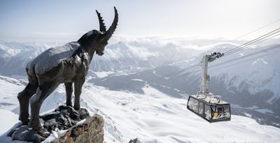 Piz Nair Winter Vorne links im Bild steht eine Steinbock-Statue des Piz Nairs. Links im Bild ist die schwebende Luftseilbahn Piz Nair zu sehen. Bei klarer Sicht fällt der Blick auf die verschneiten Hänge der Corviglia, den schneebedeckten Stazerwald im Tal sowie die gegenüberliegenden Berge.