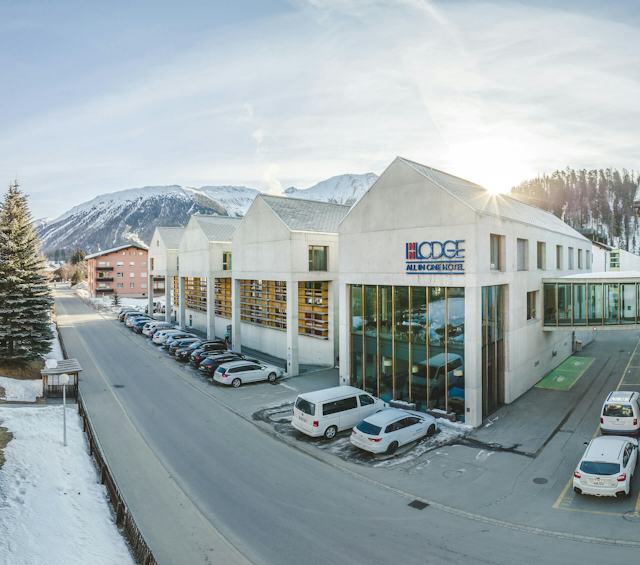 ALL IN ONE HOTEL INN LODGE Celerina | Aussenansicht Winter Links im Bild steht ein Tannenbaum. Daneben verläuft eine Strasse, auf der gegenüberliegenden Seite derer befindet sich das ALL IN ONE HOTEL INN LODGE in Celerina. Vor dem Gebäude ist ein voll belegter Parkplatz mit verschiedenen Autos. Die Wiesen und Berge sind mit Schnee bedeckt.