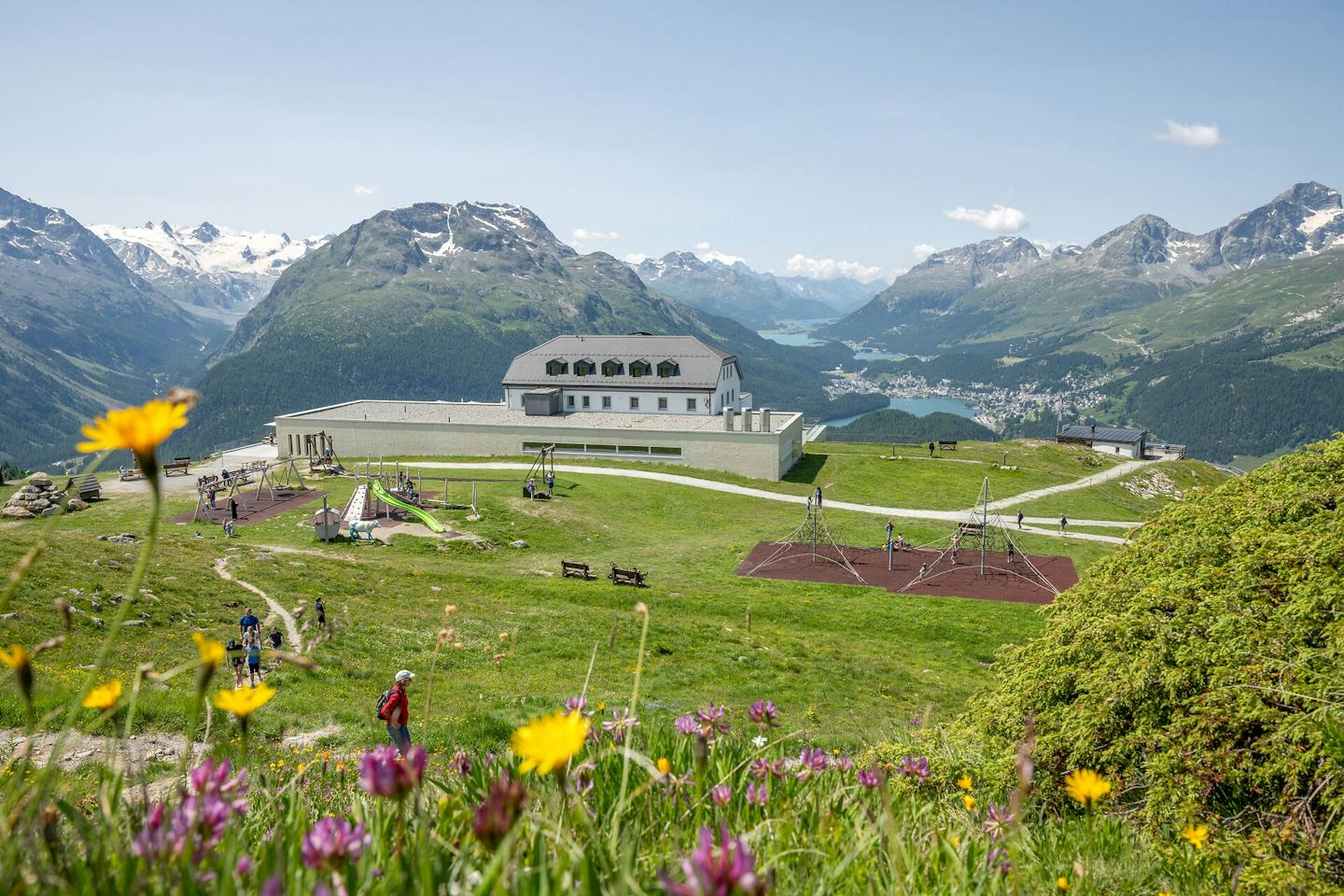 Die Berglandschaft des Engadins, gesehen von Muottas Muragl. Im Vordergrund liegt eine grüne Wiese mit gelben, roten und rosafarbenen Blumen. Im Bildmittelpunkt ist das Romantik Hotel Muottas Muragl mit Kinderspielplatz. Dahinter erstrecken sich grüne Hänge und Täler mit den Oberengadiner Seen, über denen die Gipfel der Engadiner Alpen unter einem klaren Himmel thronen.