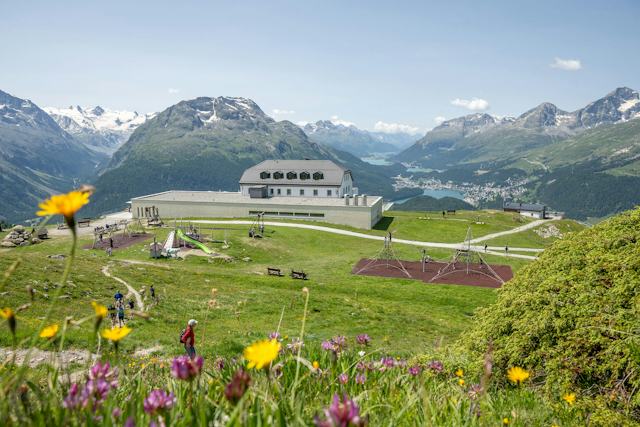 Die Berglandschaft des Engadins, gesehen von Muottas Muragl. Im Vordergrund liegt eine grüne Wiese mit gelben, roten und rosafarbenen Blumen. Im Bildmittelpunkt ist das Romantik Hotel Muottas Muragl mit Kinderspielplatz. Dahinter erstrecken sich grüne Hänge und Täler mit den Oberengadiner Seen, über denen die Gipfel der Engadiner Alpen unter einem klaren Himmel thronen.