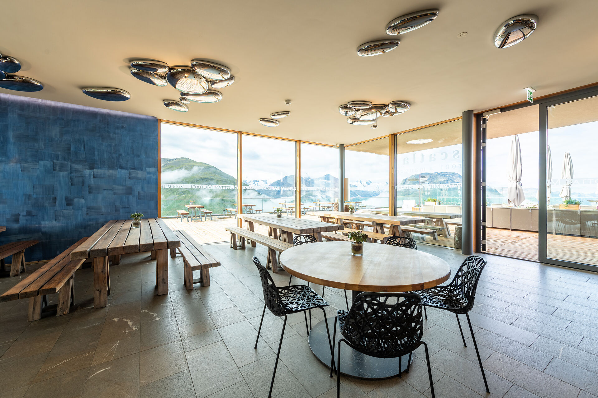 Das Restaurant Scatla – pront’ a manger auf Muottas Muragl präsentiert sich mit einem modernen, hellen Innenraum, Holztischen und einer grossen bodentiefen Fensterfront, die den Blick auf die Engadiner Bergwelt freigibt.