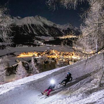 Nachtschlitteln Celerina Zwei Personen fahren nachts mit Schlitten eine verschneite, von Bäumen gesäumte Schlittelpiste oberhalb von Celerina hinunter. Eine der Personen trägt eine Stirnlampe. Im Hintergrund sind das beleuchtete Dorf Celerina und die schneebedeckten Engadiner Berge zu sehen.