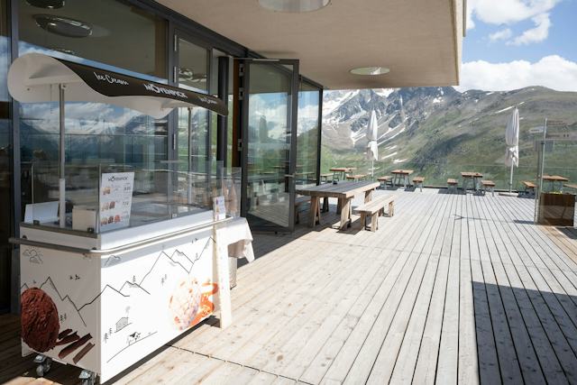 Aussenbereich des Scatla – pront’ a manger mit Eiswagen. Zahlreiche Holztischen und Sitzbänken auf einer grossen Holzterrasse. Zwei Sonnenschirme stehen geschlossen zwischen den Sitzplätzen.