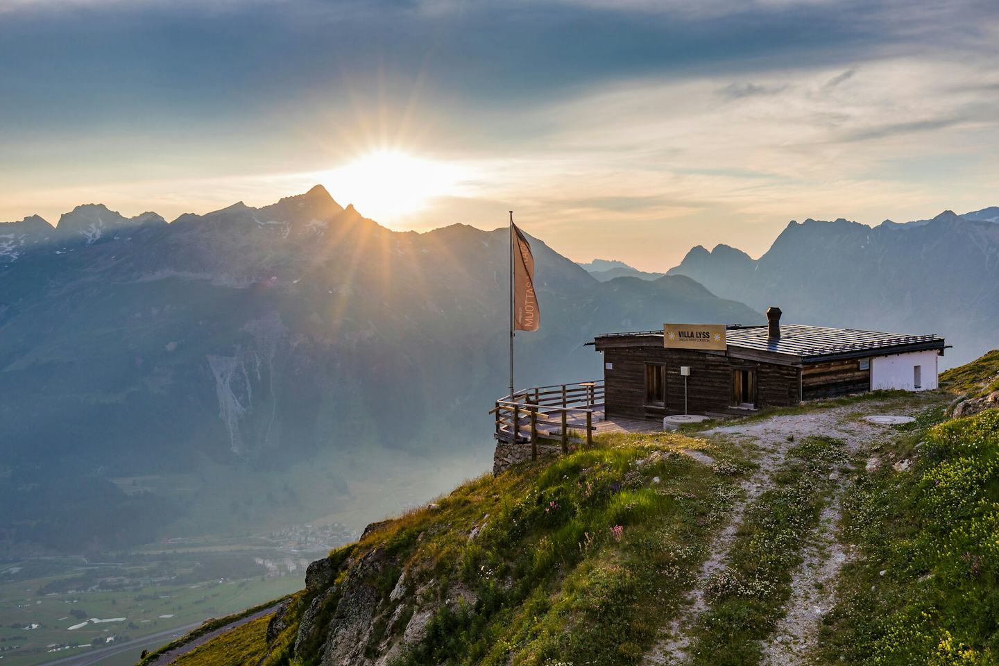 Villa Lyss | Romantik Hotel Muottas Muragl | Sommer Die Villa Lyss steht auf einem begrünten, teilweise felsigen Berghang. Das niedrige Gebäude hat eine dunkle Holzfassade, ein flaches Dach und eine kleine Terrasse mit Holzgeländer. Vor dem Gebäude stehen ein geschlossener Sonnenschirm und eine Fahne. Im Hintergrund erstreckt sich eine weitläufige alpine Berglandschaft. Über der Berglandschaft geht gerade die Sonne unter.