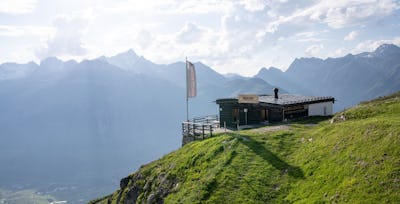 Villa Lyss | Romantik Hotel Muottas Muragl | Sommer Die Villa Lyss steht auf einem begrünten, teilweise felsigen Berghang. Das eingeschossige Gebäude hat eine dunkle Holzfassade, ein flaches Metalldach und eine kleine Terrasse mit Holzgeländer. Vor dem Gebäude steht eine Fahne. Im Hintergrund erstreckt sich eine weitläufige alpine Berglandschaft mit bewaldeten und felsigen Hängen sowie mehreren markanten, teilweise schneebedeckten Berggipfeln. Tief unterhalb des Gebäudes ist das Engadiner Tal. Über der Berglandschaft ziehen weisse und graue Wolken auf.