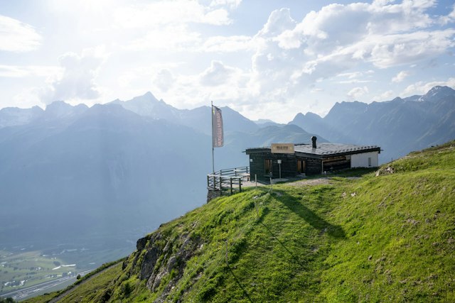 Die Villa Lyss steht auf einem begrünten, teilweise felsigen Berghang. Das eingeschossige Gebäude hat eine dunkle Holzfassade, ein flaches Metalldach und eine kleine Terrasse mit Holzgeländer. Vor dem Gebäude steht eine Fahne. Im Hintergrund erstreckt sich eine weitläufige alpine Berglandschaft mit bewaldeten und felsigen Hängen sowie mehreren markanten, teilweise schneebedeckten Berggipfeln. Tief unterhalb des Gebäudes ist das Engadiner Tal. Über der Berglandschaft ziehen weisse und graue Wolken auf.