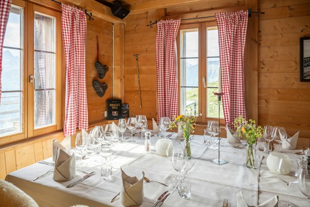 Schön eingerichteter Innenraum der Villa Lyss mit Wänden und Decke aus hellem Holz. Vor einem Fenster mit rot-weiss karierten Vorhängen steht ein grosser, weiss gedeckter Tisch. Der Tisch ist mit zahlreichen Weingläsern, Wassergläsern, Besteck, Tellern und gefalteten weissen Servietten eingedeckt. In der Mitte stehen mehrere kleine Glasvasen mit gelben und weissen Blumen sowie Kerzenständer. Um den Tisch stehen Holzbänke und Stühle mit hellen Fellauflagen. An der Holzwand hängen zwei gerahmte Bilder mit alpinen Landschaftsmotiven. Links und rechts sind Türen beziehungsweise Durchgänge sichtbar.