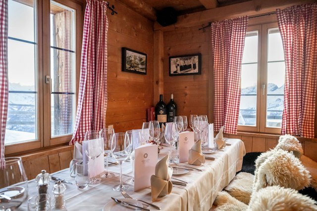 Innenraum der Villa Lyss mit Wänden und Decke aus hellem Holz. Ein langer, weiss gedeckter Tisch ist mit Tellern, Besteck, zahlreichen Wein- und Wassergläsern sowie gefalteten weissen Servietten eingedeckt. Um den Tisch stehen Holzstühle, deren Sitz- und Rückenflächen mit hellen Fellauflagen bedeckt sind. An der Holzwand hängen drei gerahmte Bilder mit Landschaftsmotiven. In der Mitte befindet sich ein Fenster mit rot-weiss karierten Vorhängen.