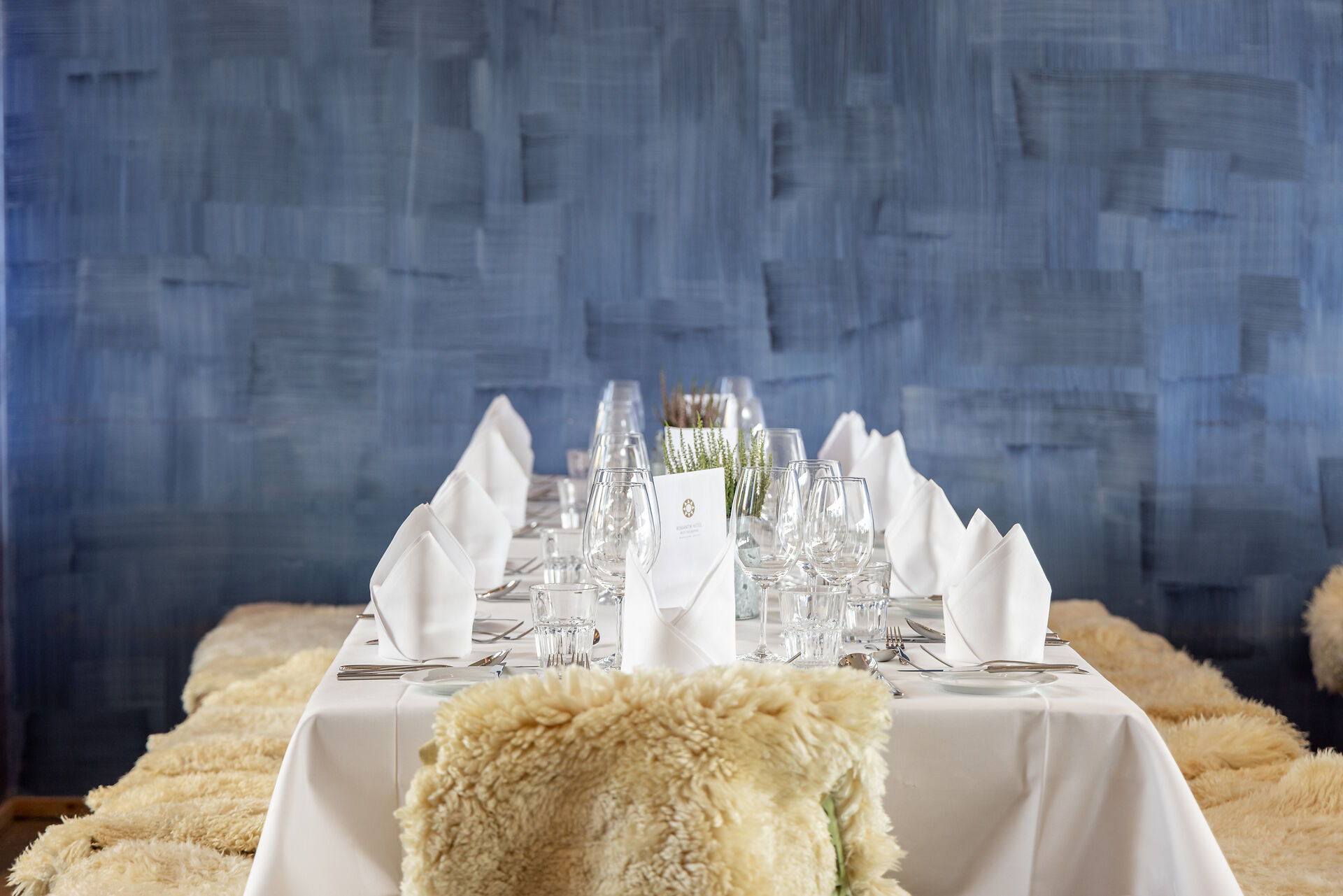 Scatla – pront’ a manger und ein langer Tischer ist mit einer weissen Tischdecke, Tellern, Besteck, Gläsern, gefalteten weissen Servietten und kleinen Pflanzen dekoriert.