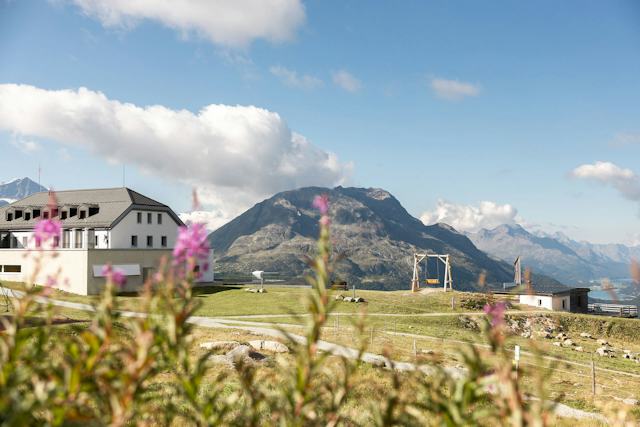 Romantik Hotel Muottas Muragl | Sommer Die Panoramaschaukel steht neben dem Gebäude des Romantik Hotel Muottas Muragl. Dahinter erstreckt sich eine weitläufige Berglandschaft mit bewaldeten Hängen, markanten Berggipfeln und einem Tal mit mehreren Seen. Im Vordergrund und seitlich der Schaukel ist eine begrünte Fläche sichtbar.