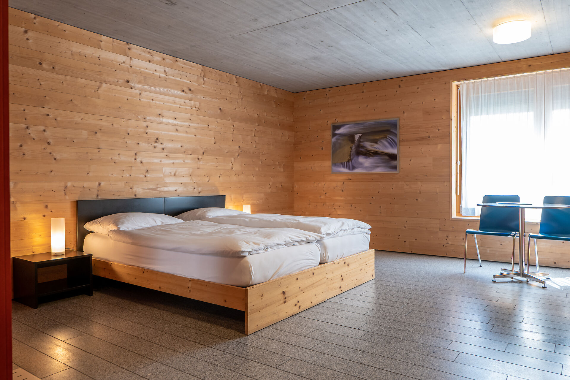 Studio S der Inn Lodge mit zwei Einzelbetten mit weisser Bettwäsche in einem Holzrahmen. Neben den Betten stehen schwarze Nachttische mit weissen Lampen. Der Raum ist vollständig mit Holz verkleidet und hat einen grauen Boden. Rechts befindet sich ein Fenster mit weissen Vorhängen sowie ein Tisch mit zwei blauen Stühlen. An der Wand hängt ein gerahmtes Bild