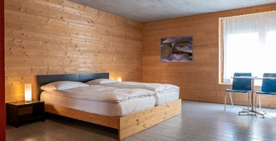 Studio S | ALL IN ONE HOTEL INN LODGE Studio S der Inn Lodge mit zwei Einzelbetten mit weisser Bettwäsche in einem Holzrahmen. Neben den Betten stehen schwarze Nachttische mit weissen Lampen. Der Raum ist vollständig mit Holz verkleidet und hat einen grauen Boden. Rechts befindet sich ein Fenster mit weissen Vorhängen sowie ein Tisch mit zwei blauen Stühlen. An der Wand hängt ein gerahmtes Bild