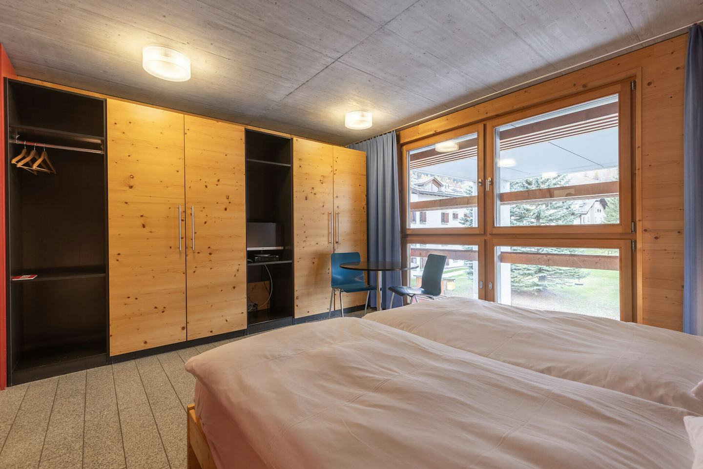 Wohn- und Schlafbereich eines Studio S der Inn Lodge mit einem Doppelbett mit weisser Bettwäsche. Im Hintergrund ist sind schränke aus Holz und rechts im Bild eine Fensterfront mit blauen Vorhängen.