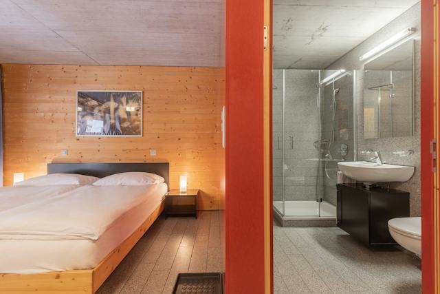 Wohn- und Schlafbereich eines Studio S der Inn Lodge mit einem Doppelbett mit weisser Bettwäsche. An der grauen Betonwand hängt ein gerahmtes Bild. Auf der rechten Seite der Blick in ein Grau gefliestes Bad, mit WC, Waschbecken und Dusche.
