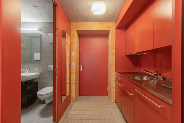 Eingangsbereich eines Studio S der Inn Lodge mit roter Eingangstür und roter Einbauküche mit Spüle, Armatur, Arbeitsfläche, Schubladen und Oberschränken. Links befindet sich ein Badezimmer mit Waschbecken, Toilette und grossem Spiegel. Die Bereiche sind durch holzverkleidete Wände und einen grauen Steinboden miteinander verbunden. Links im Bild ist das WC mit Toilette, Waschbecken und Spiegel.