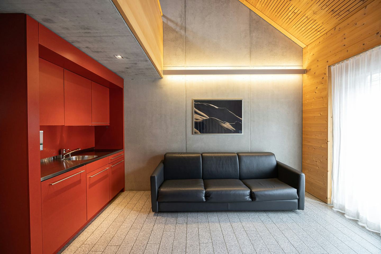 Studio M der Inn Lodge mit einem schwarzen Sofa. Im Hintergrund befindet sich eine rote Küchenzeile mit Oberschränken und Arbeitsfläche. An der grauen Wand hängt ein gerahmtes Bild.