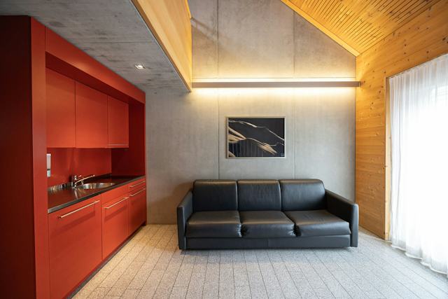 Studio M der Inn Lodge mit einem schwarzen Sofa. Im Hintergrund befindet sich eine rote Küchenzeile mit Oberschränken und Arbeitsfläche. An der grauen Wand hängt ein gerahmtes Bild.