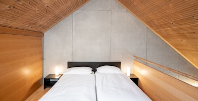 Studio M | ALL IN ONE HOTEL INN LODGE Schlafbereich eines Studio M der Inn Lodge mit einem Doppelbett mit weisser Bettwäsche. Schwarzer Nachttisch mit Stehlampe und Holzverkleidete Dachschräge.