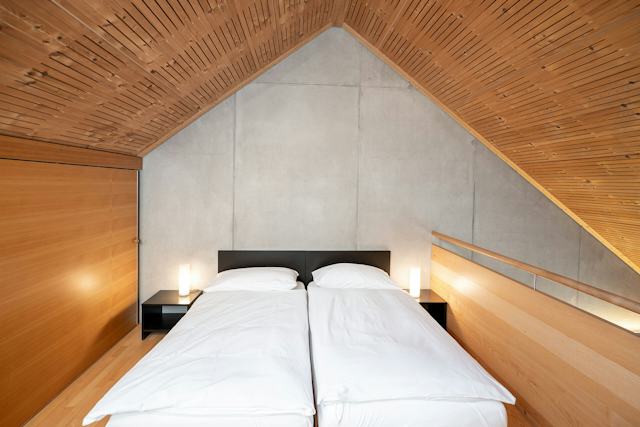 Schlafbereich eines Studio M der Inn Lodge mit einem Doppelbett mit weisser Bettwäsche. Schwarzer Nachttisch mit Stehlampe und Holzverkleidete Dachschräge.