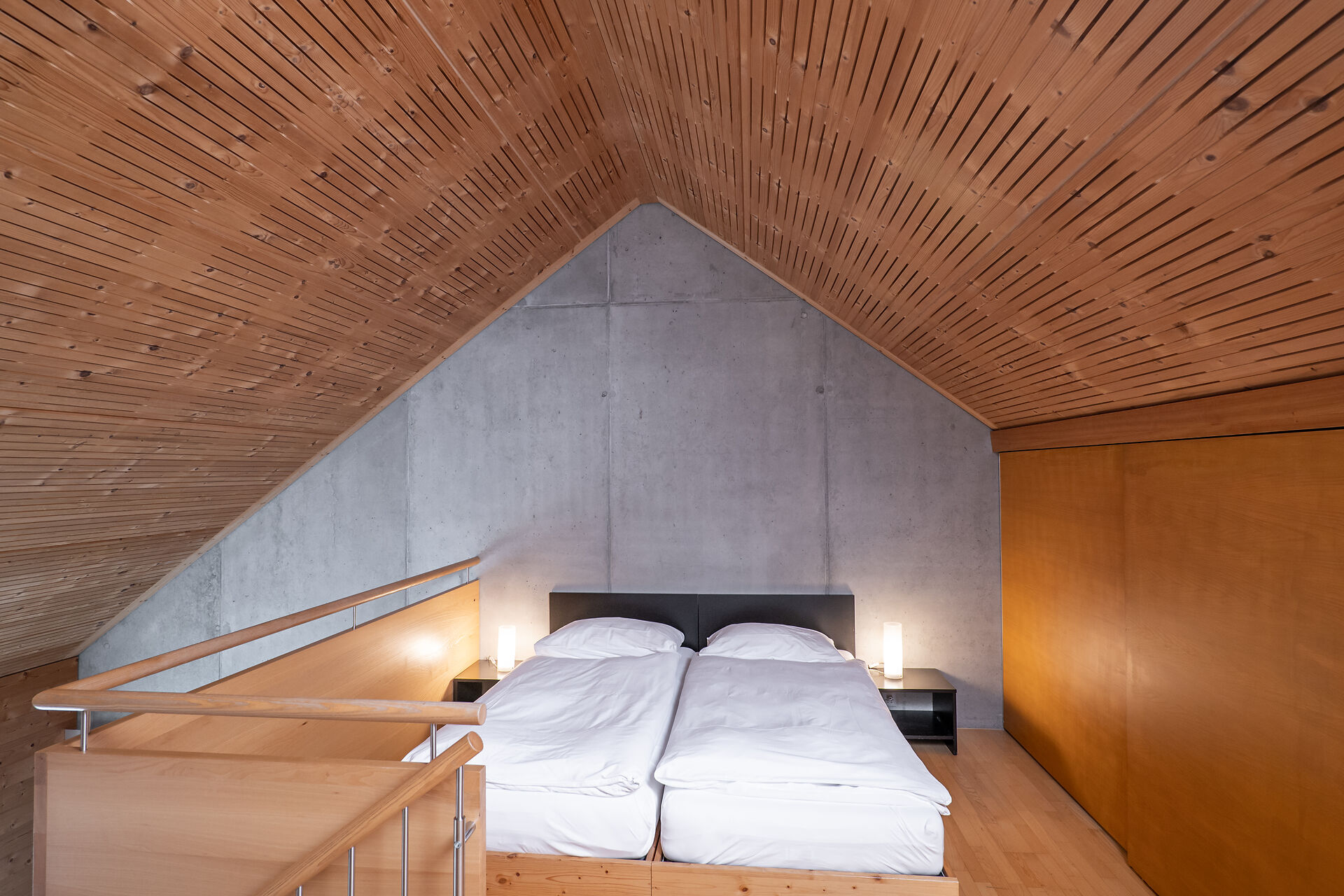 Schlafbereich eines Studio L der Inn Lodge mit zwei Einzelbetten mit weisser Bettwäsche. Die Betten stehen unter einer schrägen Holzdecke und sind durch ein Holzgeländer mit Metallstäben vom darunterliegenden Bereich abgegrenzt. Neben den Betten befinden sich schwarze Nachttische mit Lampen. An der Wand hängt ein gerahmtes Bild.