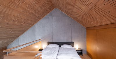 Studio L | ALL IN ONE HOTEL INN LODGE Schlafbereich eines Studio L der Inn Lodge mit zwei Einzelbetten mit weisser Bettwäsche. Die Betten stehen unter einer schrägen Holzdecke und sind durch ein Holzgeländer mit Metallstäben vom darunterliegenden Bereich abgegrenzt. Neben den Betten befinden sich schwarze Nachttische mit Lampen. An der Wand hängt ein gerahmtes Bild.