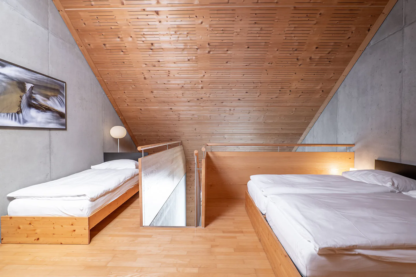 Studio L | ALL IN ONE HOTEL INN LODGE Schlafbereich eines Studio L der Inn Lodge mit zwei Betten auf einer erhöhten Holzplattform und einem weiteren Bett im Vordergrund. Die Betten sind mit weisser Bettwäsche bezogen. Der Raum verfügt über eine Holzdecke und einen Holzboden sowie eine graue Betonwand. Zwischen den Schlafbereichen befindet sich ein Holzgeländer mit Metallstäben. Neben dem Doppelbett steht ein schwarzer Nachttisch mit einer Lampe.