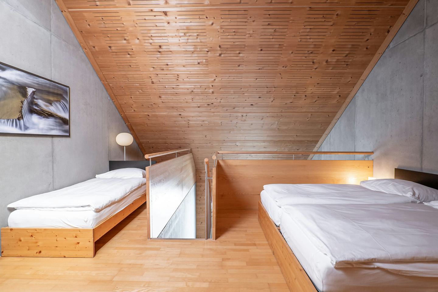 Schlafbereich eines Studio L der Inn Lodge mit zwei Betten auf einer erhöhten Holzplattform und einem weiteren Bett im Vordergrund. Die Betten sind mit weisser Bettwäsche bezogen. Der Raum verfügt über eine Holzdecke und einen Holzboden sowie eine graue Betonwand. Zwischen den Schlafbereichen befindet sich ein Holzgeländer mit Metallstäben. Neben dem Doppelbett steht ein schwarzer Nachttisch mit einer Lampe.
