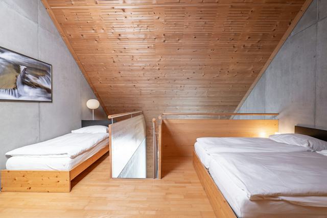 Schlafbereich eines Studio L der Inn Lodge mit zwei Betten auf einer erhöhten Holzplattform und einem weiteren Bett im Vordergrund. Die Betten sind mit weisser Bettwäsche bezogen. Der Raum verfügt über eine Holzdecke und einen Holzboden sowie eine graue Betonwand. Zwischen den Schlafbereichen befindet sich ein Holzgeländer mit Metallstäben. Neben dem Doppelbett steht ein schwarzer Nachttisch mit einer Lampe.