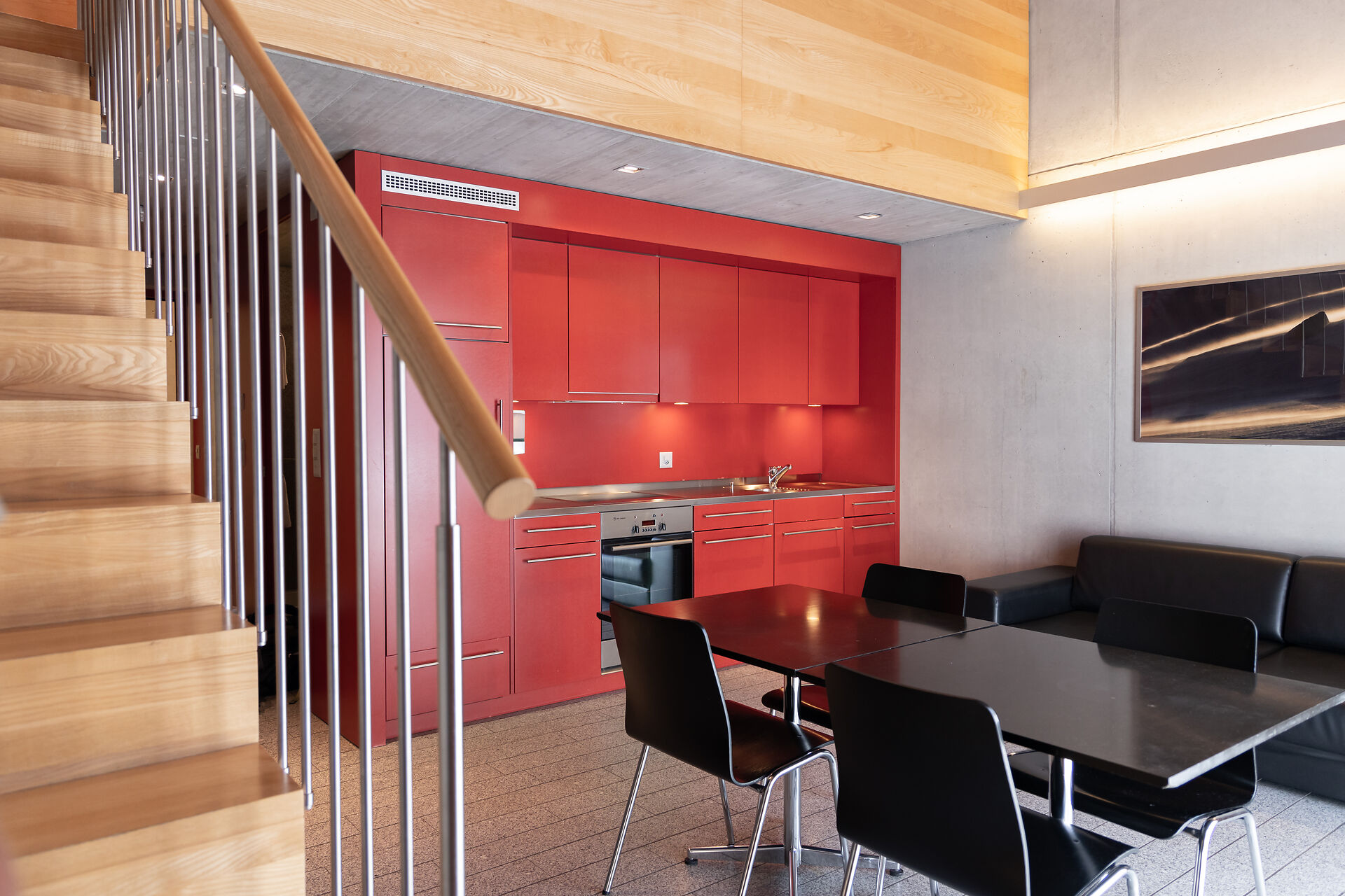 Wohnbereich eines Studio XL der Inn Lodge mit roter Einbauküche, schwarzem Esstisch und vier schwarzen Stühlen. Links führt eine Holztreppe mit Metallgeländer in den oberen Schlafbereich. An der Wand hängt ein gerahmtes Bild, darunter befindet sich eine schwarzes Ledersofa.