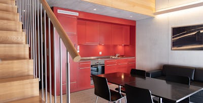 Studio XL | ALL IN ONE HOTEL INN LODGE Wohnbereich eines Studio XL der Inn Lodge mit roter Einbauküche, schwarzem Esstisch und vier schwarzen Stühlen. Links führt eine Holztreppe mit Metallgeländer in den oberen Schlafbereich. An der Wand hängt ein gerahmtes Bild, darunter befindet sich eine schwarzes Ledersofa.