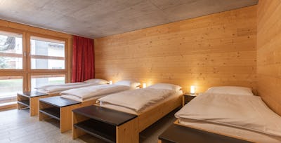 Vierbettzimmer | ALL IN ONE HOTEL INN LODGE Vierbettzimmer der Inn Lodge mit vier Einzelbetten mit weisser Bettwäsche, die in zwei Reihen angeordnet sind. Die Betten stehen in einem Raum mit holzverkleideten Wänden und Decke. An der Wand gegenüber befindet sich ein Fernseher mit roten Vorhängen.