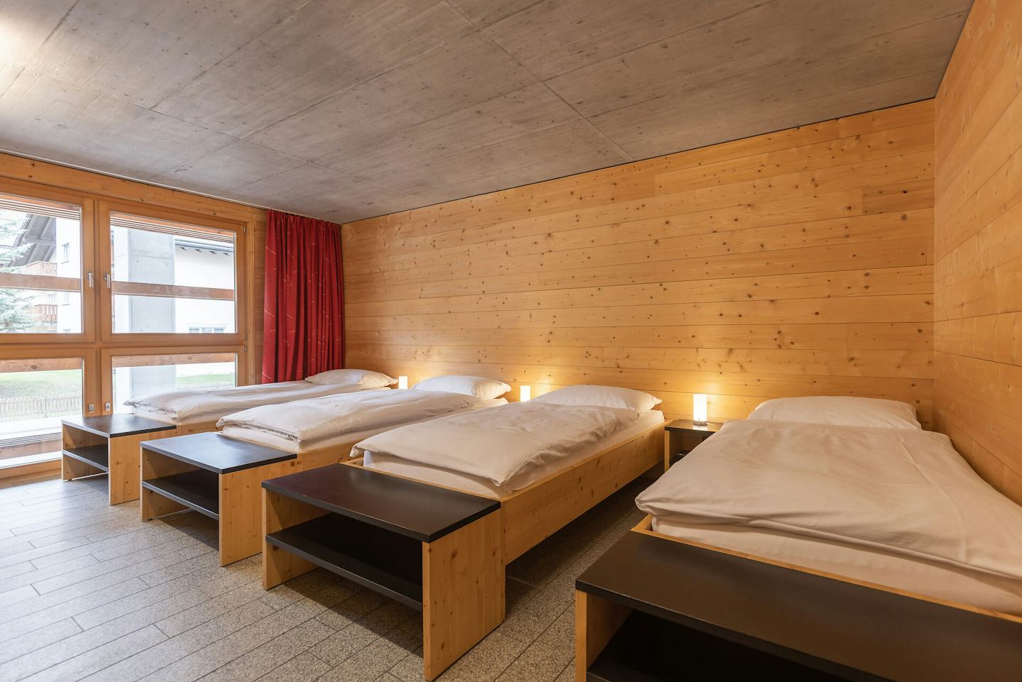 Vierbettzimmer der Inn Lodge mit vier Einzelbetten mit weisser Bettwäsche, die in zwei Reihen angeordnet sind. Die Betten stehen in einem Raum mit holzverkleideten Wänden und Decke. An der Wand gegenüber befindet sich ein Fernseher mit roten Vorhängen.