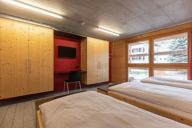 Vierbettzimmer der Inn Lodge mit vier Einzelbetten mit weisser Bettwäsche, die in zwei Reihen angeordnet sind. Die Betten stehen in einem Raum mit holzverkleideten Wänden und Decke. An der Wand gegenüber befindet sich zwei bodentiefe Fernseher.