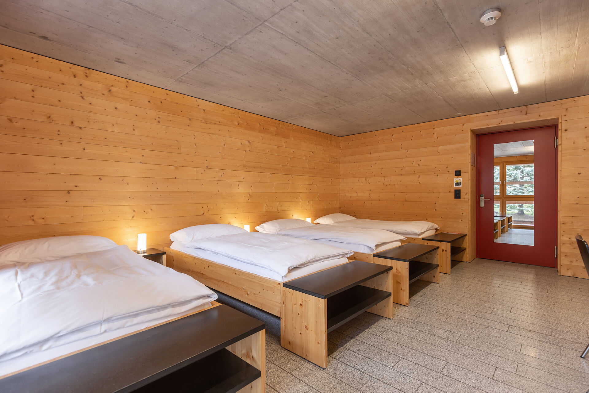 Vierbettzimmer der Inn Lodge mit vier Einzelbetten mit weisser Bettwäsche, die in zwei Reihen angeordnet sind. Die Betten stehen in einem Raum mit holzverkleideten Wänden und Decke. An der Wand gegenüber befindet sich eine rote Türe mit Spiegel. Neben den Betten stehen kleine Nachttische mit Lampen.