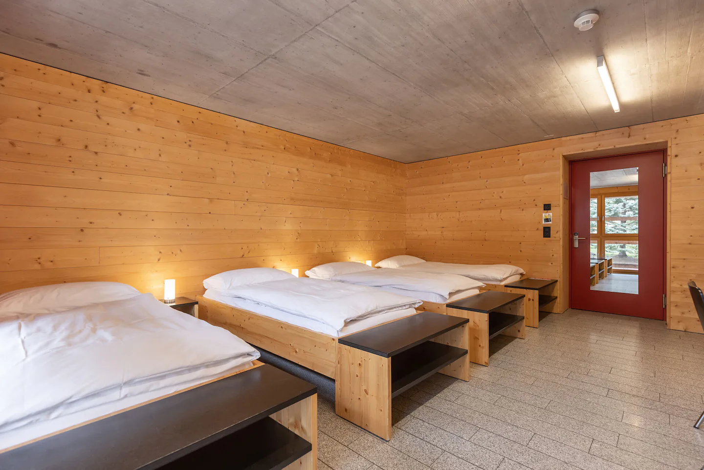 Vierbettzimmer | ALL IN ONE HOTEL INN LODGE Vierbettzimmer der Inn Lodge mit vier Einzelbetten mit weisser Bettwäsche, die in zwei Reihen angeordnet sind. Die Betten stehen in einem Raum mit holzverkleideten Wänden und Decke. An der Wand gegenüber befindet sich eine rote Türe mit Spiegel. Neben den Betten stehen kleine Nachttische mit Lampen.