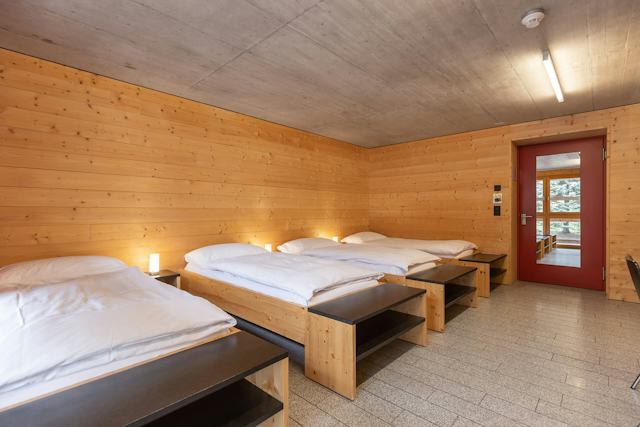 Vierbettzimmer der Inn Lodge mit vier Einzelbetten mit weisser Bettwäsche, die in zwei Reihen angeordnet sind. Die Betten stehen in einem Raum mit holzverkleideten Wänden und Decke. An der Wand gegenüber befindet sich eine rote Türe mit Spiegel. Neben den Betten stehen kleine Nachttische mit Lampen.