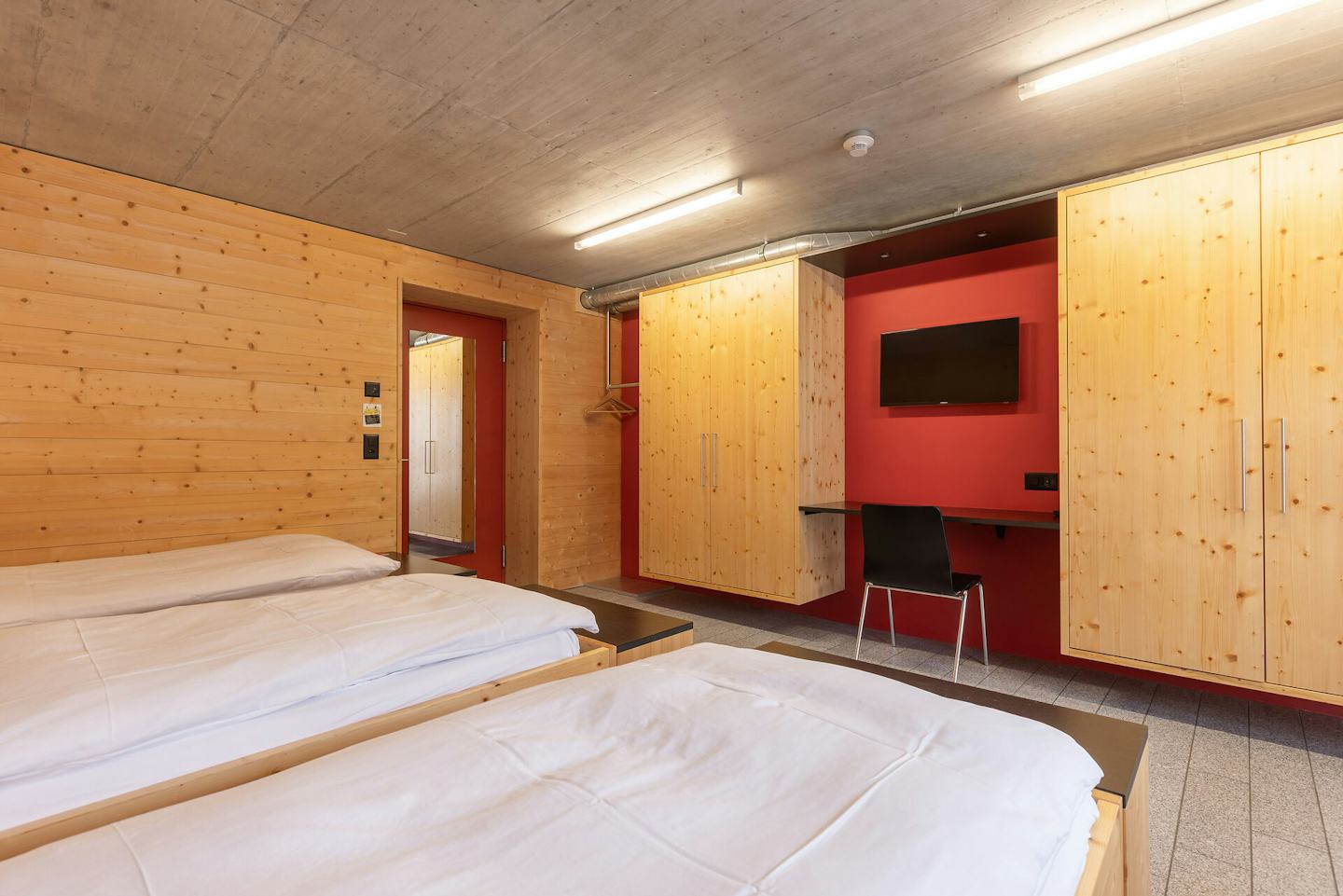 Vierbettzimmer | ALL IN ONE HOTEL INN LODGE Vierbettzimmer der Inn Lodge mit vier Einzelbetten mit weisser Bettwäsche in einer holzverkleideten Schlafnische. Gegenüber befinden sich mehrere helle Holzschränke sowie ein Arbeitsplatz mit schwarzem Stuhl und einem an der roten Wand montierten Fernseher. Im Hintergrund ist eine Tür zum Eingangsbereich zu sehen